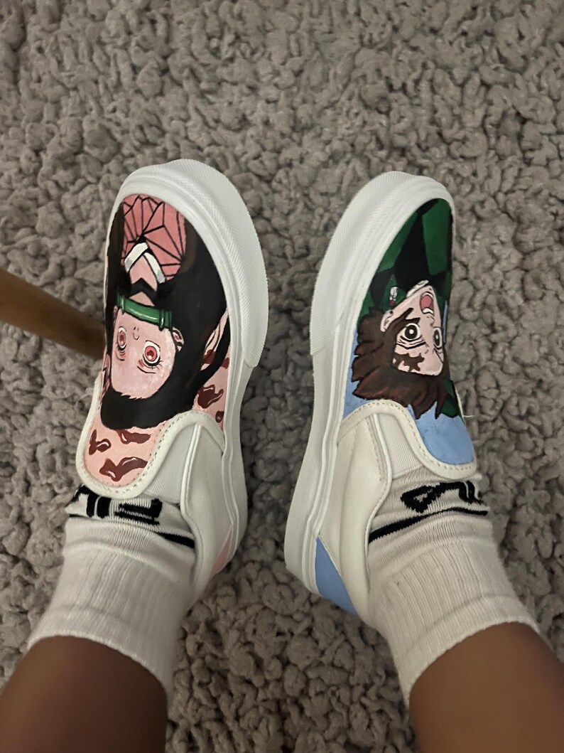 May include: White slip-on trainers, each decorated with anime character artwork. One shoe has pink accents, the other green and blue. The trainers are worn with white socks. The shoes are on a grey carpet.