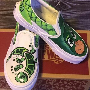 May include: Pair of white slip-on trainers with custom green character designs. One shoe features a turtle design, the other a character face. The trainers have white soles and are on a brown box.