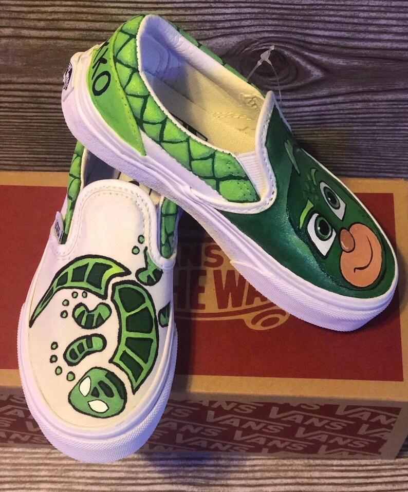 May include: Pair of white slip-on trainers with custom green character designs. One shoe features a turtle design, the other a character face. The trainers have white soles and are on a brown box.