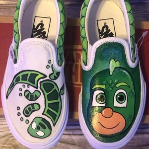 May include: White slip-on trainers with custom green artwork. One shoe has a turtle design, the other a cartoon character face. The trim and heel are green with a scale pattern.