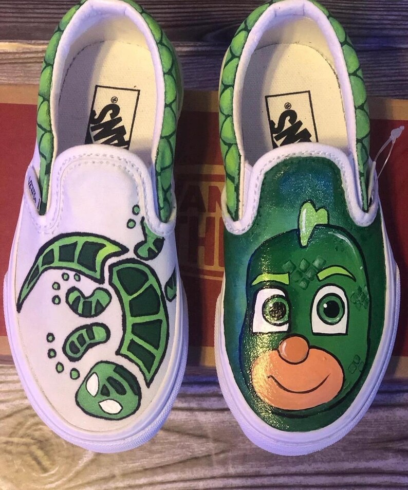 May include: White slip-on trainers with custom green artwork. One shoe has a turtle design, the other a cartoon character face. The trim and heel are green with a scale pattern.