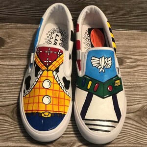 May include: Pair of white slip-on trainers, each uniquely hand-painted. One trainer features a Woody design with a yellow plaid shirt, the other a Buzz Lightyear design with a blue and green space suit. The trainers have a white sole.