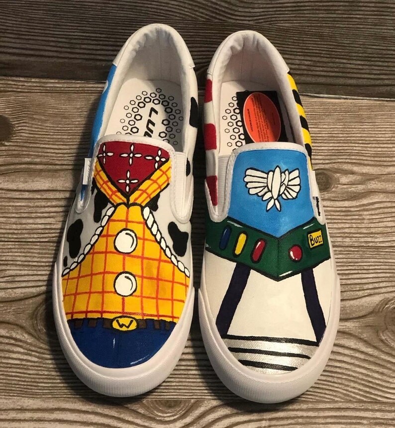 May include: Pair of white slip-on trainers, each uniquely hand-painted. One trainer features a Woody design with a yellow plaid shirt, the other a Buzz Lightyear design with a blue and green space suit. The trainers have a white sole.