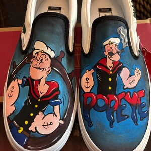 May include: Pair of white slip-on trainers with a hand-painted Popeye the Sailor design. The shoes have a black trim and a blue background with Popeye holding a ship's wheel and the word "Popeye" in red.