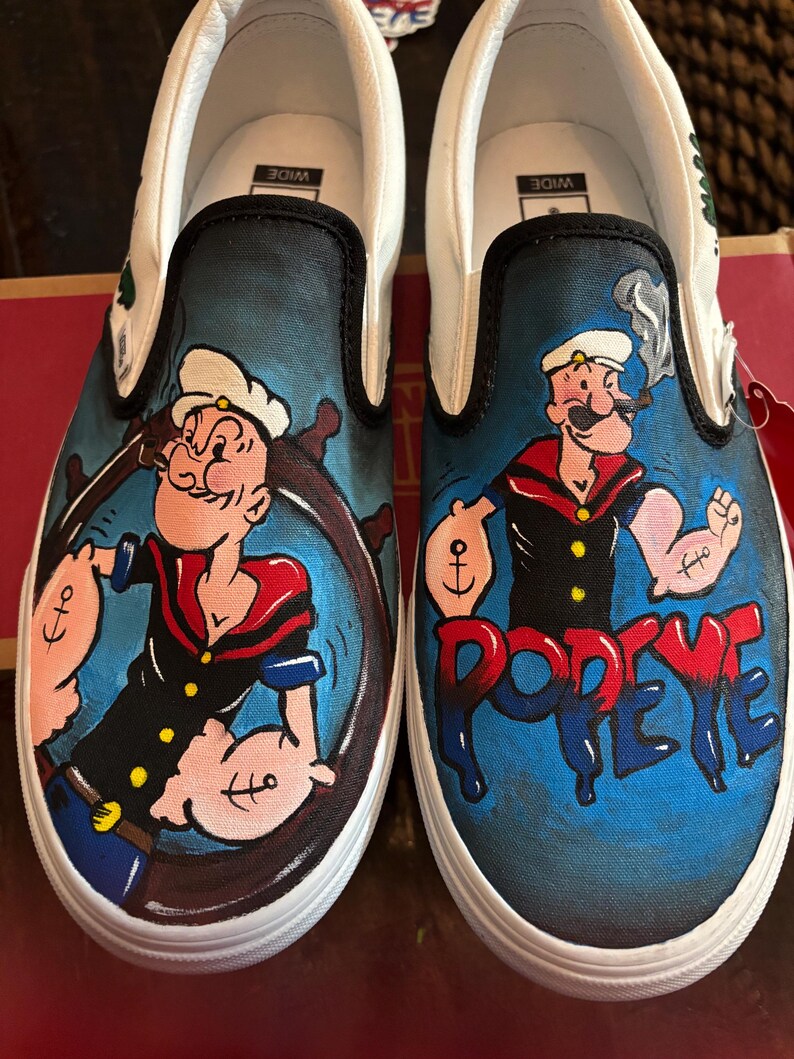 May include: Pair of white slip-on trainers with a hand-painted Popeye the Sailor design. The shoes have a black trim and a blue background with Popeye holding a ship's wheel and the word "Popeye" in red.