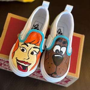May include: Pair of white slip-on trainers with cartoon character designs. One shoe depicts a character with orange hair and red lipstick, the other a brown dog. Both have blue trim and white soles.