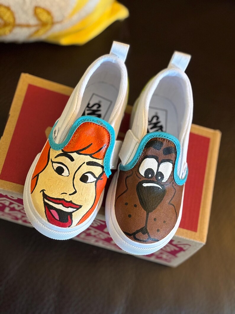 May include: Pair of white slip-on trainers with cartoon character designs. One shoe depicts a character with orange hair and red lipstick, the other a brown dog. Both have blue trim and white soles.