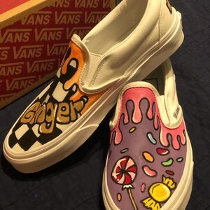 May include: White slip-on trainers with custom artwork. One shoe has the word "Ginger" in black and gold over a chequered pattern. The other features a candy design with a lollipop and dripping pink icing. The shoes are a UK size.