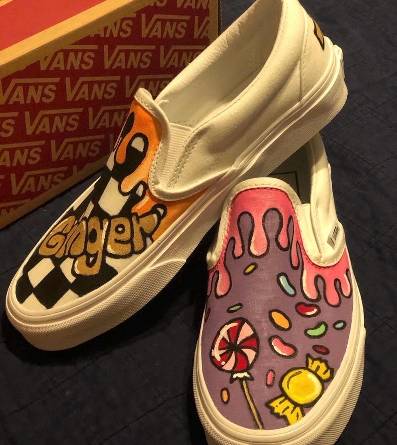 May include: White slip-on trainers with custom artwork. One shoe has the word "Ginger" in black and gold over a chequered pattern. The other features a candy design with a lollipop and dripping pink icing. The shoes are a UK size.
