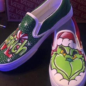 May include: Custom-painted slip-on trainers with a green and red Grinch design. One shoe features the word "Grinch" with snowflakes, the other shows the Grinch's face in a Santa hat. The trainers have white soles.