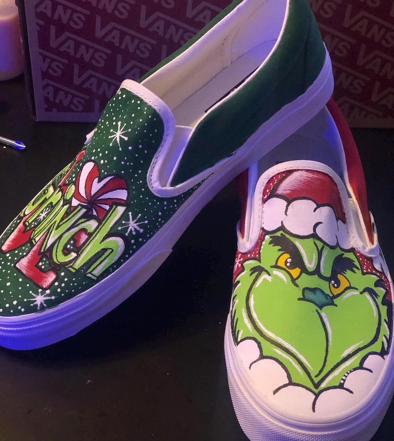May include: Custom-painted slip-on trainers with a green and red Grinch design. One shoe features the word "Grinch" with snowflakes, the other shows the Grinch's face in a Santa hat. The trainers have white soles.