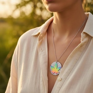 May include: Silver necklace with a circular pendant featuring a rainbow-colored cannabis leaf design. The pendant has a glittery background and is suspended from a delicate chain. The person is wearing a light-colored button-down shirt.