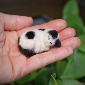 Può includere: Una figura di gatto in feltro in miniatura, fatta a mano. Il gatto addormentato è bianco con macchie nere e una testa nera. Il piccolo gatto è rannicchiato, apparendo morbido e coccoloso. La figura è tenuta nella mano di una persona.