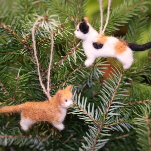 Könnte beinhalten: Zwei katzenförmige Weihnachtsschmuckstücke. Eines ist orange und weiß, das andere ist weiß mit schwarzen und orangefarbenen Flecken. Beide sind mit Schnur aufgehängt und vor einem Hintergrund aus grünen Tannennadeln platziert.