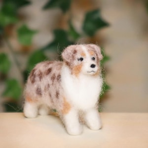 Könnte beinhalten: Handgefertigte Filzhundefigur. Der Hund hat eine weiße Brust und Beine, mit einer Mischung aus braunem, beigem und weißem Fell an Körper und Kopf. Der Hund steht auf einer hellen Oberfläche.