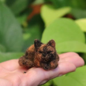 Può includere: Una piccola figurina di gatto fatta a mano in feltro, con pelo marrone, nero e arancione. Il gatto è rannicchiato in una posizione di sonno.