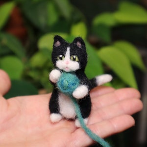 Op de afbeelding: Een kleine zwart-witte kat met groene ogen houdt een bolletje blauwe wol vast. De kat zit op de hand van een persoon.