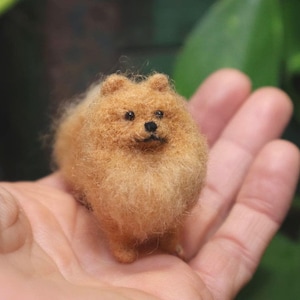Puede incluir: Una pequeña figurita de perro de fieltro de aguja, con un pelaje marrón claro. El perro está de pie sobre sus cuatro patas y tiene una cara redonda y linda.