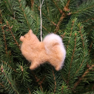 Pode incluir: Um enfeite de feltro de um cão castanho claro com cauda branca. O enfeite está pendurado em uma árvore de Natal verde.