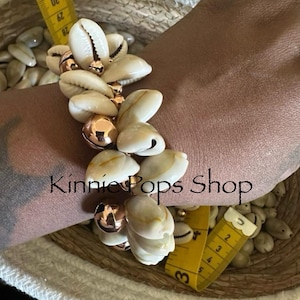 May include: A bracelet featuring natural seashells and copper-colored beads. The bracelet is adorned with multiple cowrie shells and spherical beads. The text "Kinnie Pops Shop" is visible in the image.