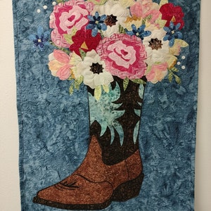 Può includere: Uno stivale da cowboy marrone con un motivo floreale blu e verde sulla parte superiore. Lo stivale è pieno di un bouquet di fiori rosa, bianchi e rossi. Lo sfondo è un tessuto blu con un motivo a spirale.
