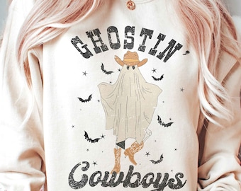 May include: A cream coloured sweatshirt with a graphic of a ghost wearing a cowboy hat and boots. The text "Ghostin' Cowboys" is printed on the sweatshirt.