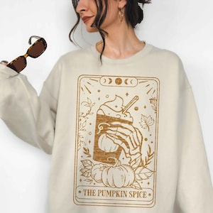 Puede incluir: Una sudadera beige con un gráfico marrón de un latte de especias de calabaza en un diseño de estilo de carta de tarot. El texto en la tarjeta dice "The Pumpkin Spice".
