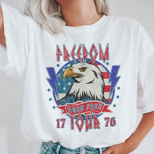 以下が含まれることがあります： アメリカの国旗と「Freedom Born Free 17 Tour 76」の文字が書かれた、ヴィンテージスタイルの鷲のグラフィックがプリントされた白いTシャツです。
