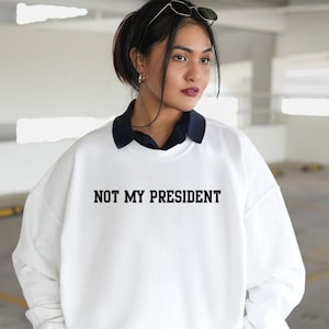 May include: White sweatshirt with the text "NOT MY PRESIDENT" in black. The sweatshirt is worn over a dark blue collared shirt and a white pleated skirt. Sunglasses rest on top of the head.