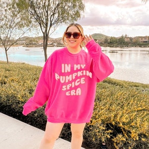 Op de afbeelding: Een vrouw draagt een roze trui met de tekst "In My Pumpkin Spice Era" in wit. Ze staat op een stoep met een meer op de achtergrond.