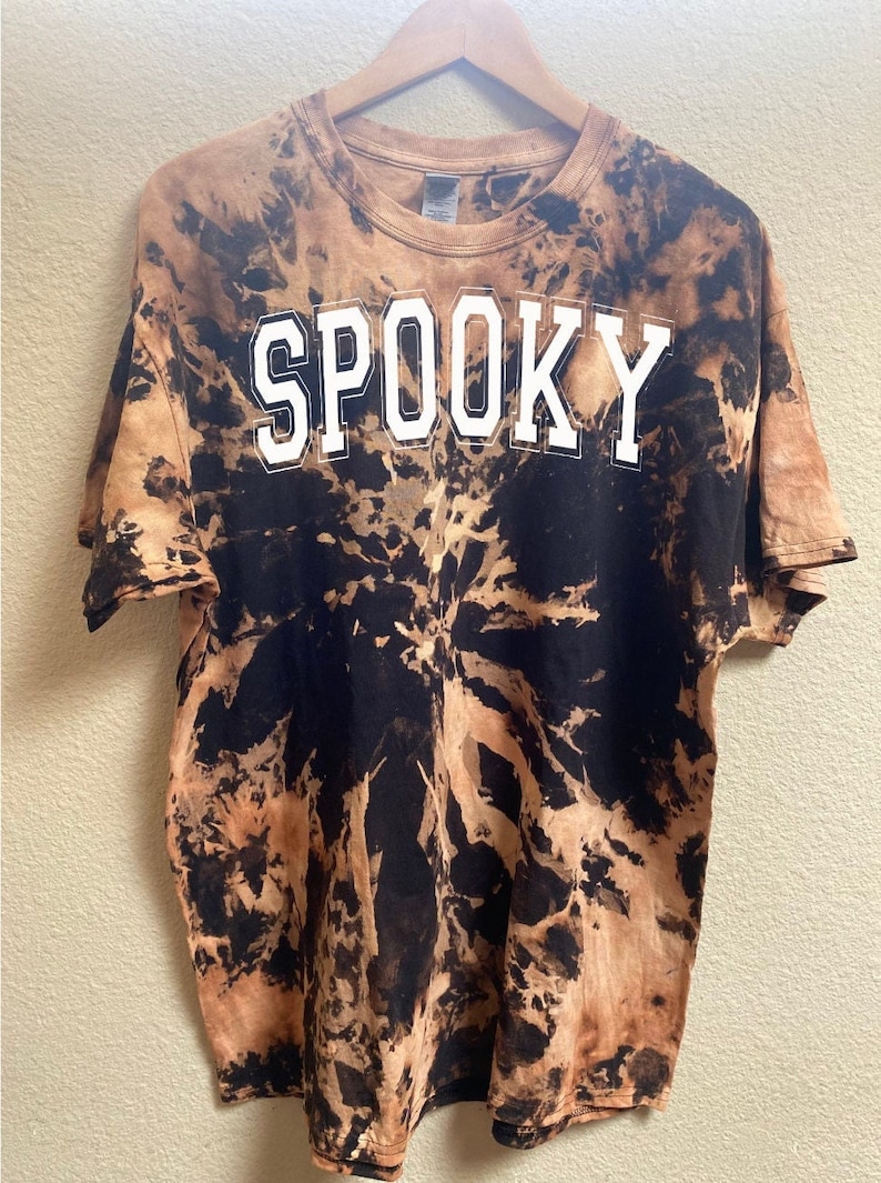 以下が含まれることがあります： 黒と茶色のタイダイ染めのTシャツで、前面に白い文字で「SPOOKY」とプリントされています。