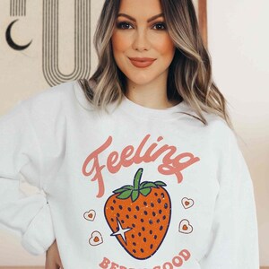 May include: White sweatshirt with a pink graphic of a strawberry and the text "Feeling Berry Good".