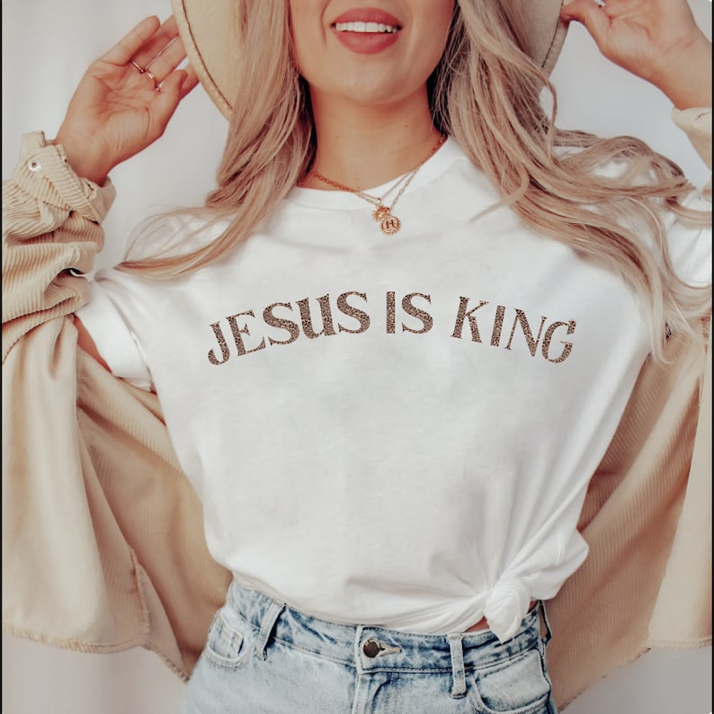 Puede incluir: Camiseta blanca con el texto "JESUS IS KING" en letras de purpurina marr&oacute;n.