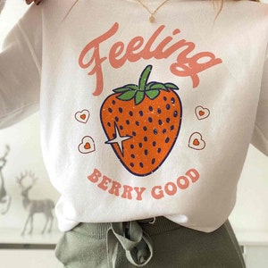 May include: White long-sleeve sweatshirt with a pink "Feeling Berry Good" graphic featuring a red strawberry with green leaves and a sparkling star.