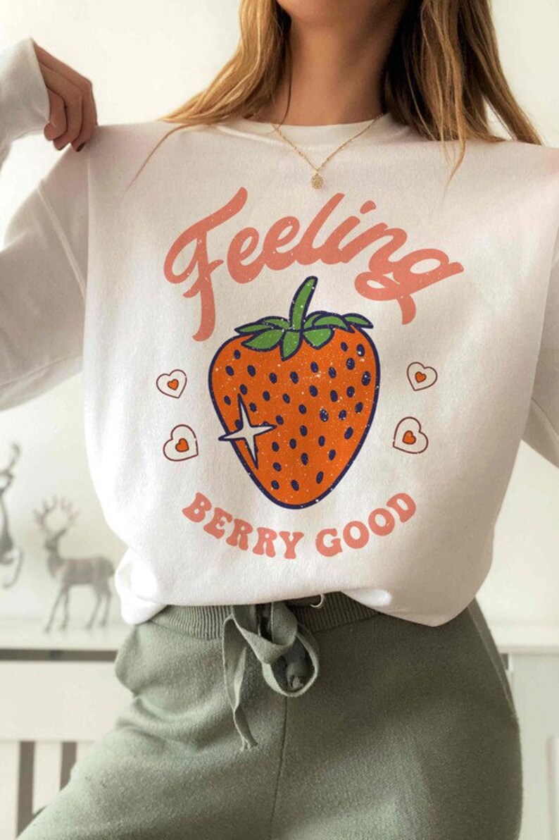 May include: White long-sleeve sweatshirt with a pink "Feeling Berry Good" graphic featuring a red strawberry with green leaves and a sparkling star.