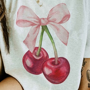 May include: A white t-shirt with a pink bow and two red cherries printed on the front. The cherries have green stems and leaves.