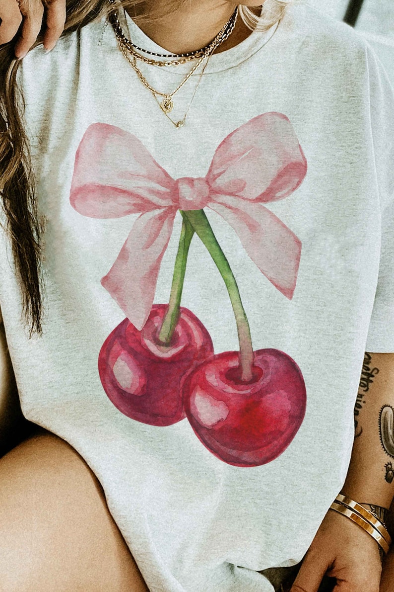 May include: A white t-shirt with a pink bow and two red cherries printed on the front. The cherries have green stems and leaves.