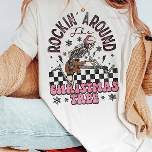K&ouml;nnte beinhalten: Wei&szlig;es T-Shirt mit einem Aufdruck eines Skeletts, das Gitarre spielt, mit dem Text "Rockin' Around the Christmas Tree" in Pink und Schwarz.