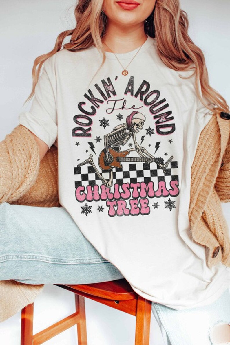K&ouml;nnte beinhalten: Wei&szlig;es T-Shirt mit einem Aufdruck eines Skeletts, das Gitarre spielt, mit dem Text "Rockin' Around the Christmas Tree" in Pink und Schwarz.