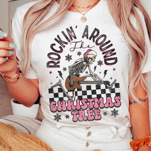 K&ouml;nnte beinhalten: Wei&szlig;es T-Shirt mit einem Grafikdruck eines Skeletts, das Gitarre spielt, mit dem Text "Rockin' Around the Christmas Tree" in Pink und Schwarz.