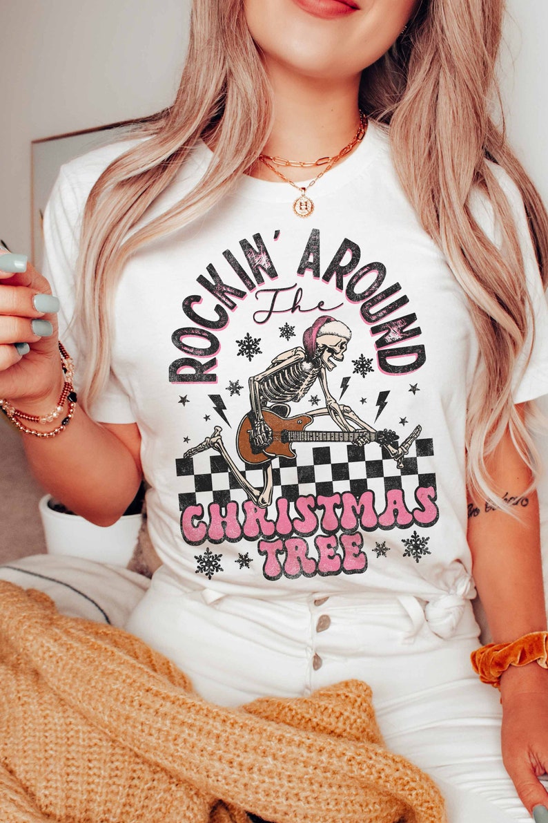 K&ouml;nnte beinhalten: Wei&szlig;es T-Shirt mit einem Grafikdruck eines Skeletts, das Gitarre spielt, mit dem Text "Rockin' Around the Christmas Tree" in Pink und Schwarz.