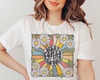 May include: White t-shirt with a retro graphic print of a sun with rainbow rays and daisies. The text "Here Comes The Sun" is printed in a groovy font.