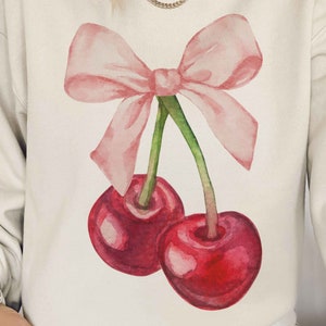 May include: A cream-colored sweatshirt with a watercolor illustration of two red cherries with green stems and a pink bow.