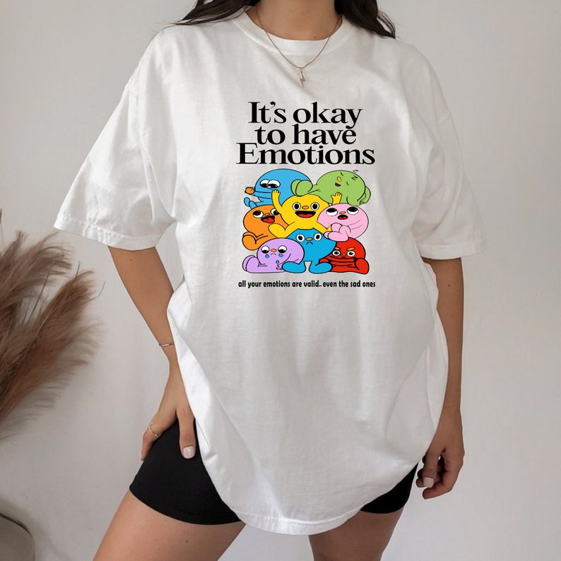 Op de afbeelding: Wit T-shirt met een afbeelding van kleurrijke cartoonfiguren en de tekst "It's okay to have Emotions" en "all your emotions are valid. even the sad ones."