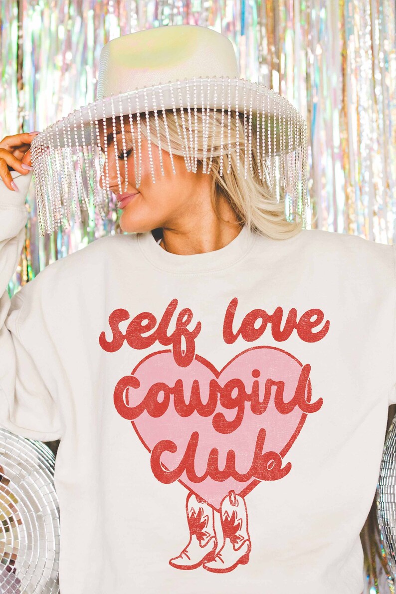 K&ouml;nnte beinhalten: Ein wei&szlig;er Pullover mit einem rosa Herz und rotem Text, der "self love cowgirl club" lautet. Das Herz hat rote Cowboystiefel unten.