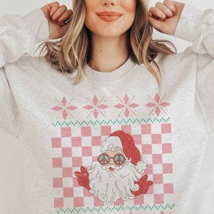 K&ouml;nnte beinhalten: Ein grauer Pullover mit einem pinkfarbenen und gr&uuml;nen Weihnachtsmotiv, das einen Cartoon-Weihnachtsmann mit Peace-Zeichen-Sonnenbrille zeigt. Das Design beinhaltet ein kariertes Muster und Schneeflocken.