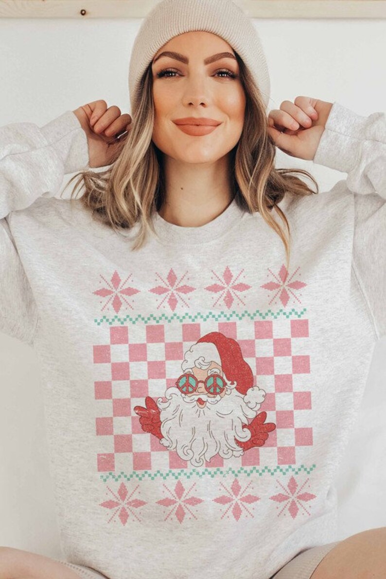 K&ouml;nnte beinhalten: Ein grauer Pullover mit einem pinkfarbenen und gr&uuml;nen Weihnachtsmotiv, das einen Cartoon-Weihnachtsmann mit Peace-Zeichen-Sonnenbrille zeigt. Das Design beinhaltet ein kariertes Muster und Schneeflocken.