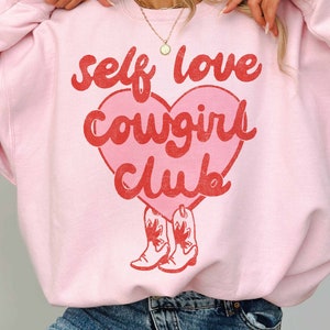 K&ouml;nnte beinhalten: Rosafarbener Pullover mit einem roten Herz-Motiv, auf dem "self love cowgirl club" steht, darunter zwei rote Cowboystiefel.
