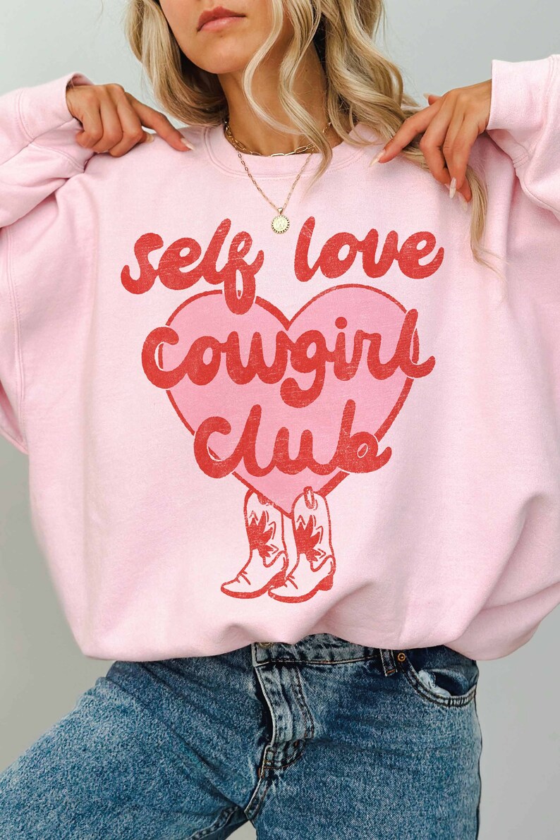 K&ouml;nnte beinhalten: Rosafarbener Pullover mit einem roten Herz-Motiv, auf dem "self love cowgirl club" steht, darunter zwei rote Cowboystiefel.