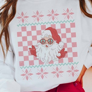 K&ouml;nnte beinhalten: Wei&szlig;er Weihnachtspullover mit einem rosa-gr&uuml;nen Karomuster, Schneeflocken und einem Retro-Weihnachtsmann mit Peace-Zeichen-Sonnenbrille.